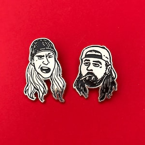 Op de afbeelding: Twee emaille pins met zwart-wit illustraties van personages met lang haar en hoeden. Eén pin toont een persoon met een open mond, de andere een persoon met een baard en een baseball cap. De pins staan tegen een rode achtergrond.