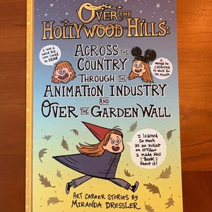 Op de afbeelding: Een boek getiteld "Over the Hollywood Hills: Across the Country Through the Animation Industry and Over the Garden Wall" met cartoon illustraties. De cover heeft tekst en de naam van de auteur. Het boek ligt op een houten oppervlak.
