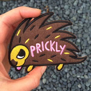 Peut inclure: Patch brodé en feutre marron représentant un hérisson de dessin animé avec des yeux jaunes et blancs, une langue rose qui sort et le mot "PRICKLY" en lettres roses.