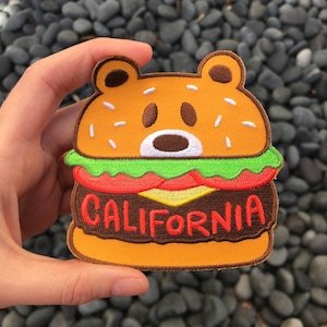 May include: A brown bear-shaped embroidered patch in the shape of a hamburger. The hamburger has a green lettuce, red tomato, yellow cheese, and a brown patty. The words "CALIFORNIA" are embroidered in red on the patty.