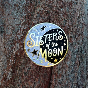 以下が含まれることがあります： 三日月と星が描かれた金と黒のエナメルピン。"Sisters of the Moon"というテキストが、遊び心のあるフォントで書かれています。