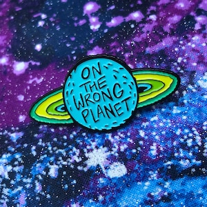 May include: A blue enamel pin featuring a planet with rings. The planet has the text "ON THE WRONG PLANET" on it.