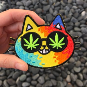 May include: A colorful embroidered patch of a cat wearing sunglasses with a marijuana leaf design. The cat is yellow, blue, green, and orange.