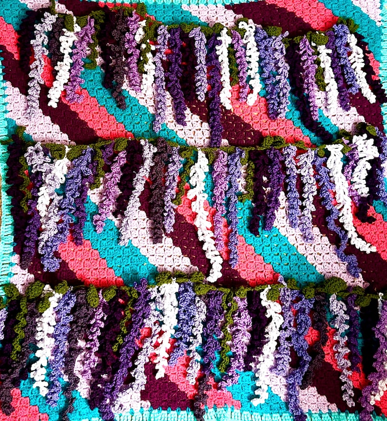 K&ouml;nnte beinhalten: Eine handgefertigte H&auml;keldecke mit farbenfrohem Design. Die Decke hat einen t&uuml;rkisfarbenen, rosa- und lilafarbenen Hintergrund mit kaskadenartigen, glyzinienartigen floralen Akzenten in Lila-, Wei&szlig;- und Gr&uuml;nt&ouml;nen. Die Decke ist ein gem&uuml;tlicher, dekorativer Artikel.
