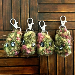 May include: Four handmade keychains with a fuzzy, textured appearance. The keychains feature a mix of green, pink, and gold yarn, with large, cartoonish eyes. Each keychain has a silver-coloured clasp for attaching to keys or bags.