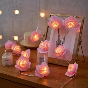May include: A garland of pink rose fairy lights, each with a warm white glow. The lights are displayed on a wooden surface, alongside a framed picture and a candle. The rose lights are a decorative item.