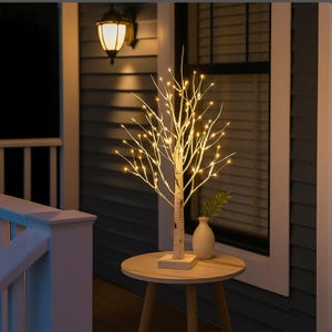 Puede incluir: Un abedul decorativo con luces LED blancas cálidas. El árbol está sobre una pequeña mesa redonda con un jarrón blanco y una planta verde. El fondo incluye un exterior de casa azul oscuro y una lámpara de pared.