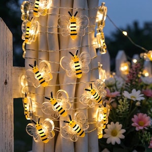 May include: Decorative string lights with bee-shaped ornaments. The bees are made of clear plastic with yellow and black stripes. The lights are strung on a thin wire and emit a warm glow. The lights are draped over a white fence and curtains.