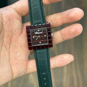Könnte beinhalten: Eine Chopard-Armbanduhr mit einer quadratischen, tiefroten, facettierten Lünette und einem passenden Zifferblatt mit Gittermuster sowie vier kleinen Diamant-Stundenmarkierungen. Die Uhr ist an einem dunkelgrünen Lederarmband befestigt.