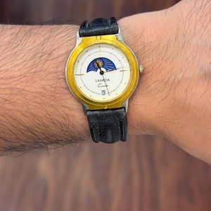 May include: A wristwatch with a black leather strap and a gold-tone bezel. The white dial features a sun and moon phase indicator at the top, black hour and minute hands, and the text LAMEDA Quartz above a date window displaying the number 3.