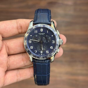 May include: A Victorinox Swiss Army chronograph watch with a dark blue dial, silver-tone hands, and Arabic numeral hour markers. The watch features a tachymeter bezel, three sub-dials, a date window at the six o'clock position, and a blue leather strap.