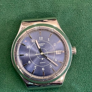 Puede incluir: Un reloj de pulsera plateado con una esfera circular azul con efecto rayos de sol, números arábigos blancos del 1 al 12, manecillas plateadas, una ventana de fecha en la posición de las 3 y el texto Swatch Swiss Automatic.
