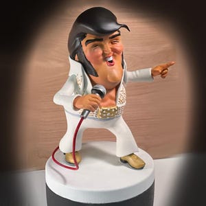 May include: A figurine of a singing performer in a white jumpsuit with gold accents, a wide gold belt, and black hair. The figurine is holding a microphone and pointing with one hand. The base is white and black.