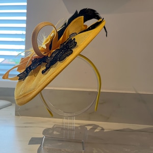 Op de afbeelding: Een gele hoed met een brede rand, versierd met zwart kant, veren en een decoratieve lus. De hoed wordt gepresenteerd op een transparante standaard, die het ontwerp benadrukt. Geschikt voor een speciale gelegenheid.