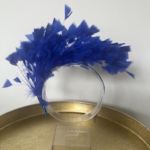 Pode incluir: Uma vibrante tiara de penas azuis. A tiara apresenta um design circular adornado com um aglomerado de penas azuis brilhantes. As penas estão dispostas em forma de leque. A tiara é exibida em um suporte de plástico transparente.
