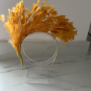 May include: A golden yellow feather headband, displayed on a clear acrylic stand. The headband has a semi-circular design with a gold-coloured band and a large cluster of feathers on one side. The feathers are a warm, vibrant yellow.