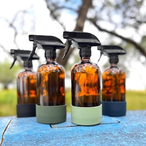 May include: Four amber glass spray bottles with black spray nozzles and colored silicone bases. The bases are in shades of green and blue. The bottles are arranged on a blue wooden surface.