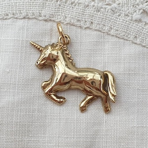 May include: A polished gold-tone metal unicorn pendant featuring a detailed mane, a single spiralled horn, and a small circular jump ring at the top for attachment. The unicorn is depicted in a prancing pose with its front legs raised and tail flowing.