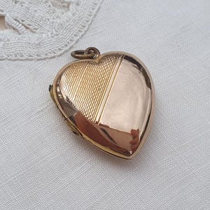 Op de afbeelding: Een hartvormige medaillonhanger in goudkleur met een verdeeld ontwerp. De ene helft van het hartoppervlak heeft een fijn, getextureerd kruisarceringpatroon, terwijl de andere helft glad en gepolijst is. Aan de bovenkant zit een klein metalen oogje.