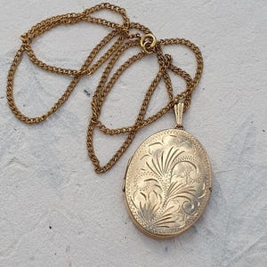 May include: An oval gold-toned locket pendant suspended from a matching gold-toned metal chain. The front surface of the locket features an intricate, engraved floral pattern with swirling lines and leaf-like motifs across the entire face.