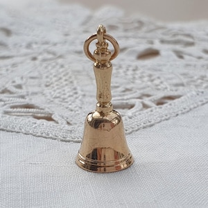 May include: A small, polished gold-tone metal bell charm featuring a slender handle and a circular jump ring at the top. The bell has a smooth, reflective surface with a flared base and a distinct, rounded top section, designed as a jewellery pendant.