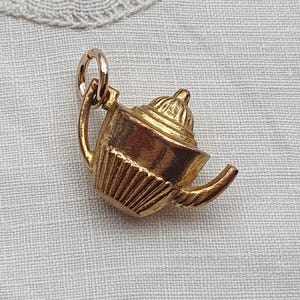 May include: A gold-toned metal charm shaped like a miniature teapot. The charm features a rounded body with vertical ribbed detailing, a curved handle, a spout, and a domed lid. A small circular jump ring is attached to the top of the handle.