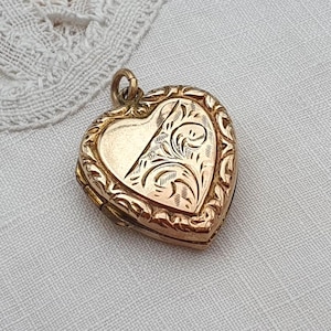 Könnte beinhalten: Ein goldfarbener herzförmiger Medaillon-Anhänger mit einem komplizierten gravierten Blumenmuster auf einer Seite und einer glatten, schlichten Oberfläche auf der anderen. Das Medaillon ist von einem dekorativen Rand umgeben und hat eine kleine Metallöse.