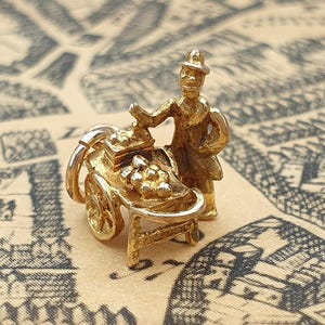 May include: A gold charm in the shape of a street vendor standing beside a small cart. The vendor wears a hat and coat, while the cart features detailed wheels and a display of small, round items. A circular jump ring is attached to the side.