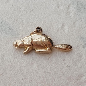 May include: A gold-toned metal charm shaped like a beaver in a crouching position, featuring a textured tail and a small circular jump ring attached to the top for suspension. The surface of the charm is smooth and reflective.