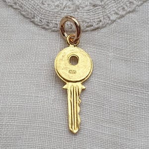 May include: A gold-coloured metal key charm featuring a circular head with a central hole and a vertical shaft with ridges. The charm includes a small jump ring at the top for attachment and a clearly visible hallmark stamp reading 375 on the head.