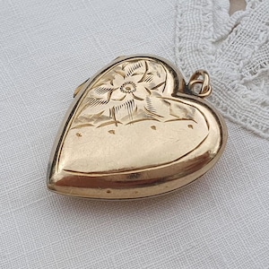 Könnte beinhalten: Ein goldfarbenes herzförmiges Medaillon mit einem gravierten Blumenmuster im oberen Bereich und einer glatten, polierten Oberfläche in der unteren Hälfte. Das Medaillon verfügt über eine kleine Metallöse an der Oberseite zur Befestigung an einer Kette.