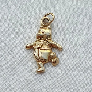 May include: A polished gold-tone metal charm shaped like a standing bear character with a textured shirt detail and a small circular jump ring attached at the top for hanging. The bear is depicted in a cheerful pose with arms outstretched.