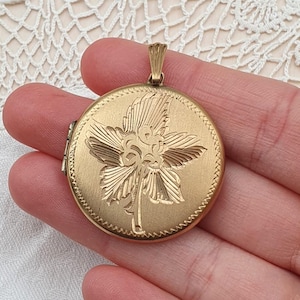 May include: A round, gold-colored locket with a floral design. The locket has a detailed flower engraving on the front and a decorative edge. It features a small loop at the top for a chain. The locket is held in a hand.