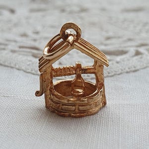 May include: A gold-coloured metal charm shaped like a miniature wishing well. The charm features a circular base with a textured stone-like appearance, a roof structure supported by two posts, and a small bucket suspended from a central horizontal bar.