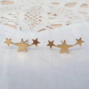 May include: A pair of gold-colored star-shaped earrings. Each earring features three stars connected by a delicate bar. The earrings are displayed on a white, textured surface.