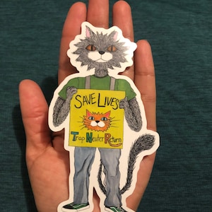 May include: A gray cat wearing a green shirt and holding a yellow sign that says "Save Lives Trap Neuter Return". The sign has a picture of an orange cat on it. The cat is wearing blue jeans and green sneakers.