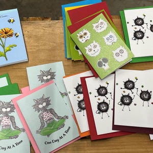 May include: Assortment of greeting cards with various designs. Some cards feature cats with different expressions, while others have a cat doing yoga with the text "One Day At A Time".  One card has a bee and flower design with the text "Just Bee Cause".