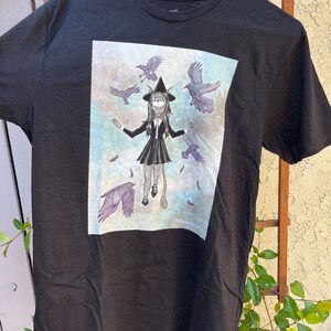 May include: Black t-shirt with a graphic print of a woman with cat ears and a witch's hat surrounded by crows. The text "Waterberry721" is printed in the bottom right corner of the graphic.