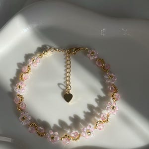 May include: A delicate bracelet featuring pink flower-shaped beads and gold-coloured accents. The bracelet has a gold-coloured chain with an adjustable length and a small heart charm. The jewellery is displayed on a white surface.