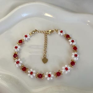 Può includere: Un braccialetto di perline fatto a mano con perline bianche a forma di margherita e perline di cristallo rosse, collegate da maglie di metallo dorato. Il braccialetto ha una catena regolabile con un ciondolo a forma di cuore. "Beads & Beyond".