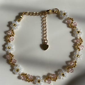 May include: A delicate gold-toned bracelet featuring a floral design. The bracelet is adorned with white and pink beads, creating a daisy-like pattern. A small heart charm dangles from the adjustable chain.