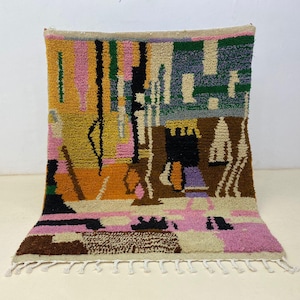 May include: A rectangular hand-knotted wool rug featuring a vibrant abstract pattern in shades of mustard yellow, pink, brown, black, green, and cream. The rug has a thick, textured pile and is finished with a row of white fringe along the bottom edge.