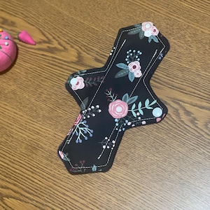 May include: A reusable menstrual pad with a black background and a floral pattern in pink, white, and green. The pad has a stitched border and a unique shape. A pink pincushion and floral arrangement are in the background.