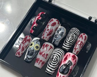 May include: A set of coffin-shaped false nails with various horror-themed designs. The designs include blood splatters, a face, a spiral, and the text "ART was here". The nails are displayed in a black case.