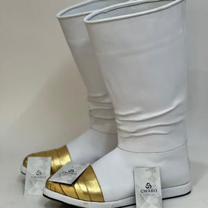 Puede incluir: Un par de botas blancas con punteras doradas. Las botas tienen una caña alta y una suela plana. Cada bota tiene una etiqueta que dice "GWABO CLOSEST".