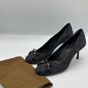 Könnte beinhalten: Ein Paar schwarze Pumps mit hohem Absatz und sich wiederholendem Logomuster. Die Schuhe haben kleine Schleifen mit Bambus-Akzenten und Quasten. Ein brauner Staubbeutel liegt neben den Schuhen.