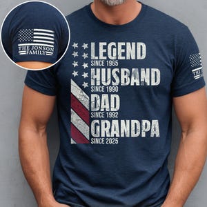 May include: Navy blue t-shirt with white text: "LEGEND SINCE 1965, HUSBAND SINCE 1990, DAD SINCE 1992, GRANDPA SINCE 2025." An American flag design is on the front. The sleeve has an American flag and "THE JONSON FAMILY".