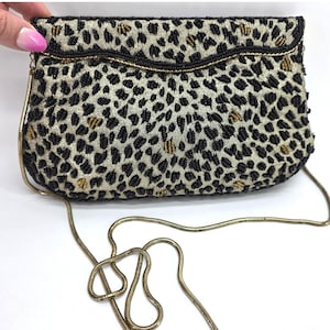 May include: A rectangular clutch bag featuring a gold fabric base with a black and gold beaded leopard print pattern. The bag has a curved flap closure and a long, thin gold-toned metal snake chain shoulder strap attached to the sides.