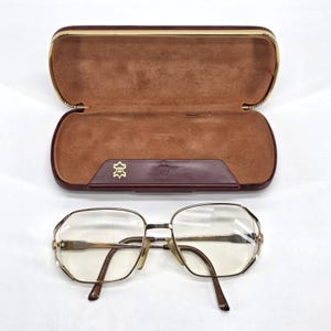 May include: Gold-toned metal eyeglasses with clear lenses and brown temple tips, positioned in front of an open, rectangular, dark burgundy hard-shell glasses case with a soft tan interior lining and a gold-colored metal trim along the edge.