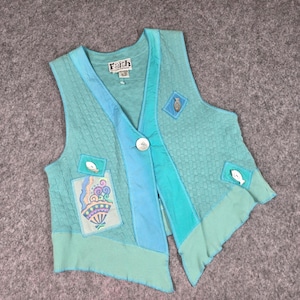 May include: A turquoise vest with a quilted pattern and a V-neck design. The vest features patches with fish and floral designs, a large white button, and the brand name "Faith". The vest has a unique, asymmetrical hemline.